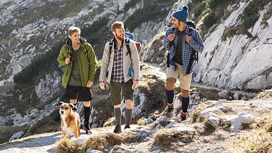 Juzo Adventure Three men with a dog in the mountains. The men are wearing compression stockings on their lower legs.