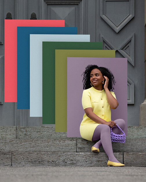 Una mujer con medias de compresión en color Cozy Violet está sentada en unas escaleras. Al fondo, los paneles de color de la actual colección Juzo Trend Colours.