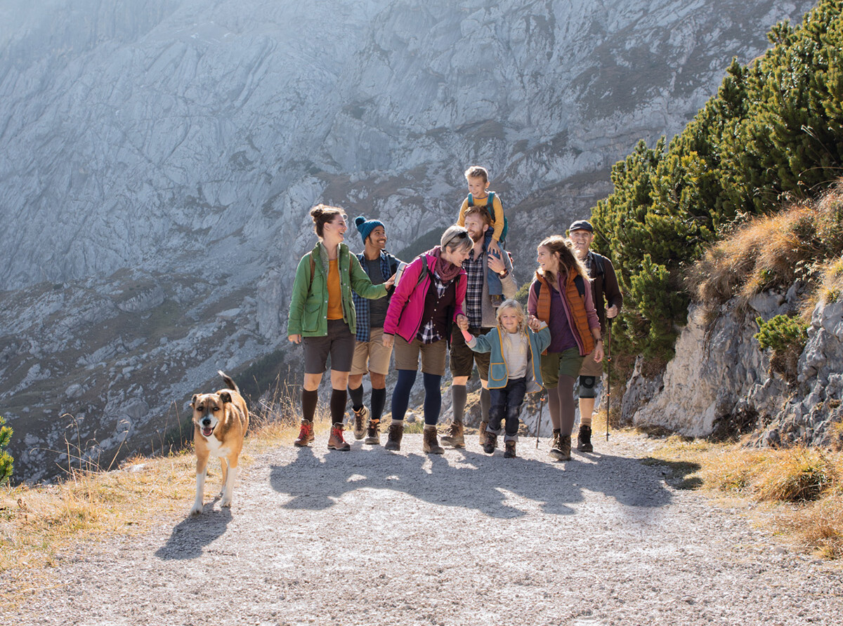 Gruppe von Wandernden auf einem Bergweg, einige mit Kompressionsstrümpfen und Bandagen, begleitet von einem Hund vor felsiger Kulisse.